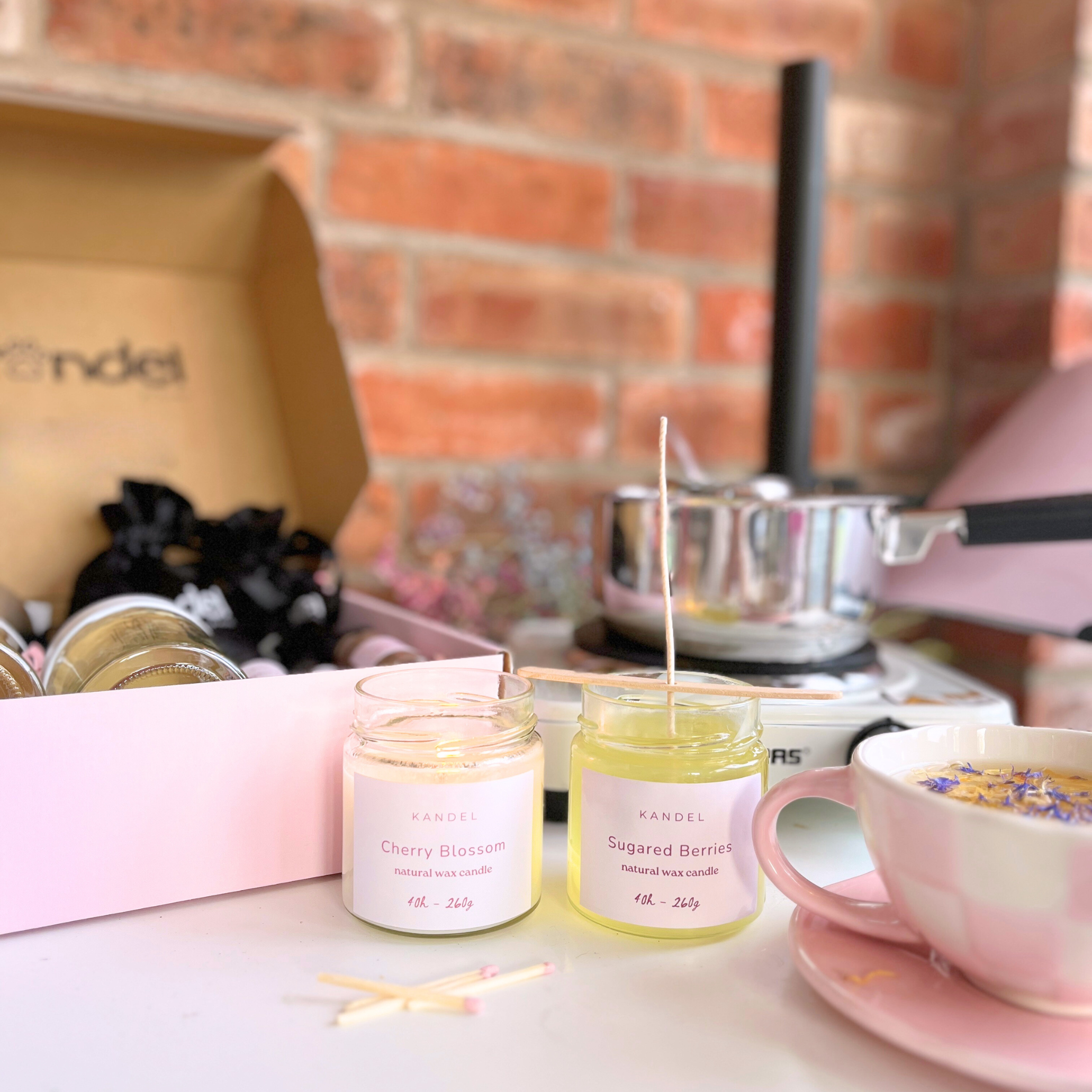 Flat lay of a candle-making kit featuring ingredients for sugared berries and cherry blossom-scented candles, including wax, wicks, fragrance oils, and containers. The setup is complemented by pink boxing in the background, adding a charming and elegant touch.