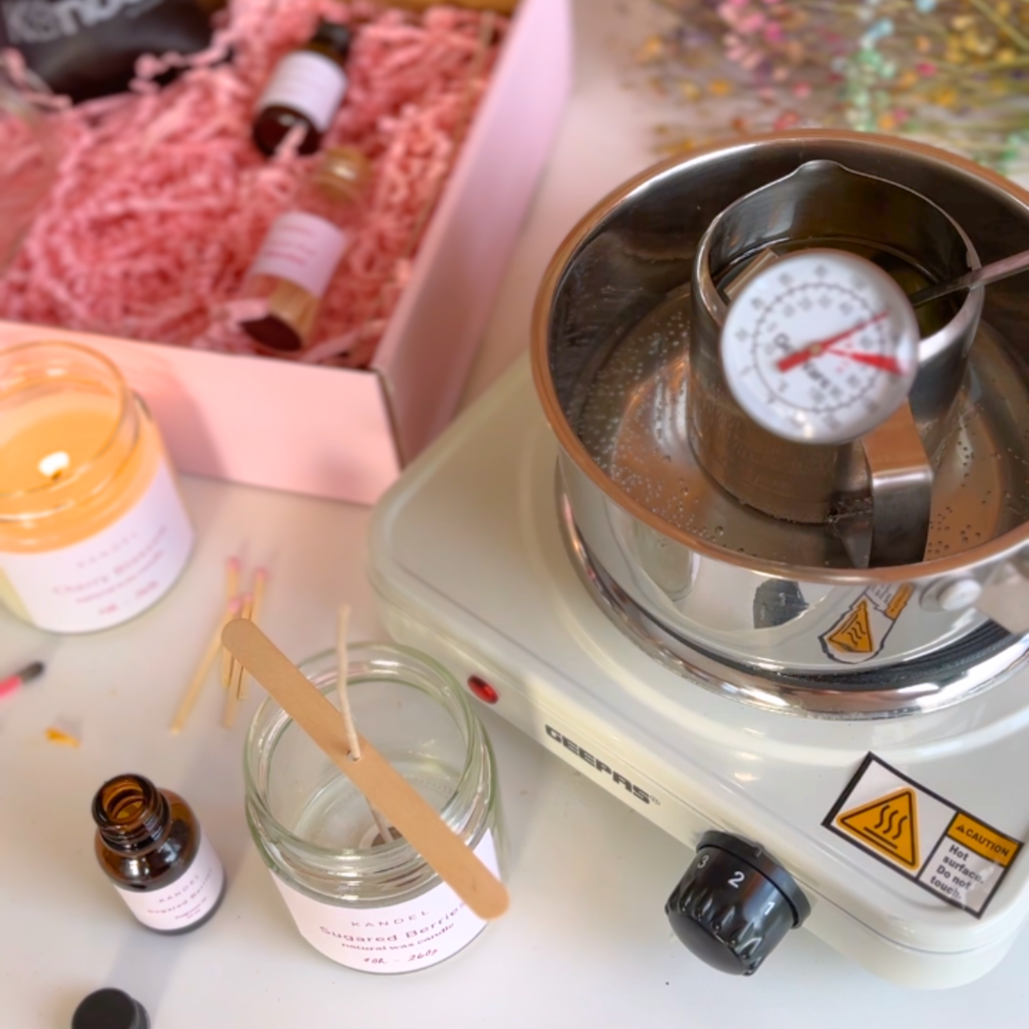 Image showing wax being boiled in a metal boiler, with a pink aesthetic background featuring a box of candle-making ingredients and fragrance oils. The setup creates a warm, inviting atmosphere for the candle-making process.